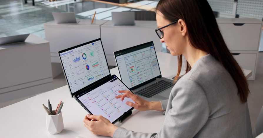 A woman working across a tablet and laptops showing a compliance dashboard